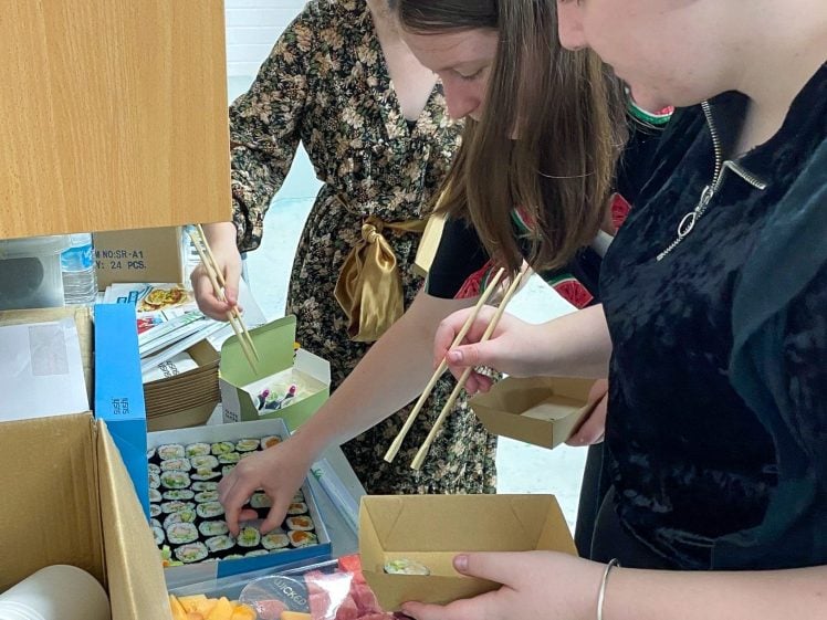 Image of Three people stand at a table using chopsticks to pick up sushi from a platter. There are also trays of sliced fruit, boxes, and other food items on the table. One person is wearing a pink beanie.