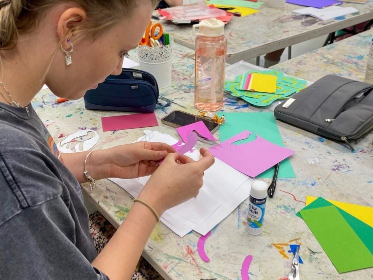 Image of Three students sit at a table in an art classroom, working with colourful paper, scissors, and glue on creative projects. The foreground shows a girl focused on cutting and assembling paper shapes.