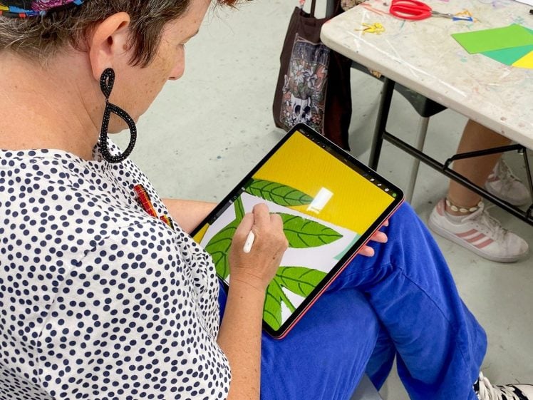 Image of A person sitting on a chair draws green leaves on a tablet with a stylus. They wear a colourful headband, polka-dot shirt, and blue trousers. Another person at a nearby table works with craft supplies.