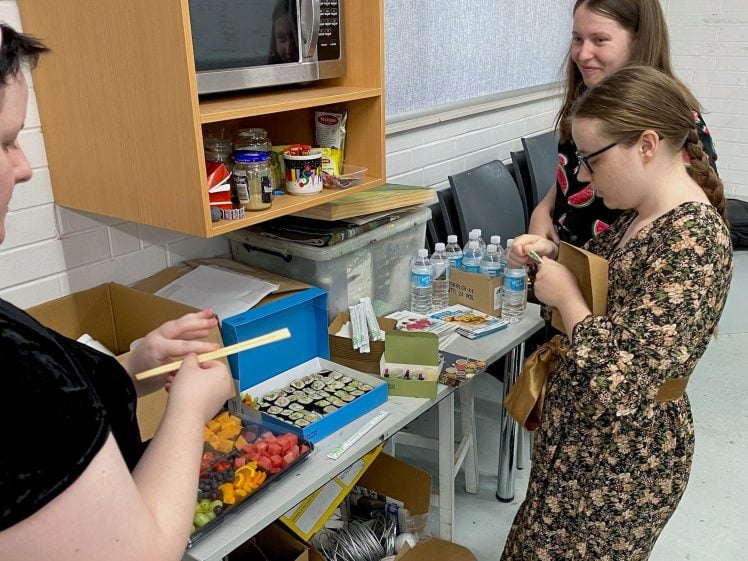 Image of Three women stand by a table with sushi, fruit, bottled water, and snacks. One woman uses chopsticks near a box of sushi, while the others watch, one holding a small plate. Kitchen items and supplies are on shelves behind them.