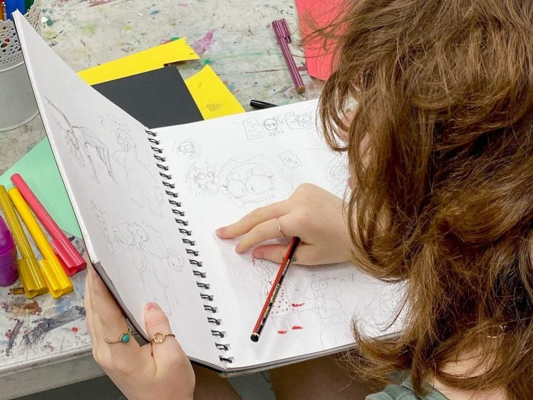 Image of A person with wavy brown hair sketching cartoon characters in a large spiral notebook, surrounded by coloured markers and paper on a messy art table.