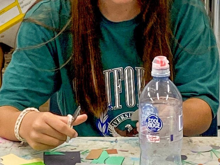 Image of A young person with long brown hair cuts coloured paper shapes at a table covered with craft materials. A bottle of water sits on the table in the foreground.