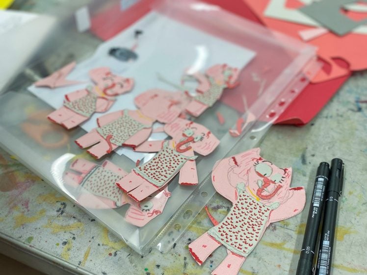 Image of Several pink paper figures with cheerful expressions are scattered on a desk, alongside black markers, paper scraps, and a clear folder on a messy, paint-stained surface.