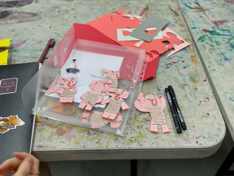 Image of A plastic folder on a messy table holds several handmade pink paper elephants, with red and grey paper, markers, and craft supplies scattered around. A person's hand rests near the folder.