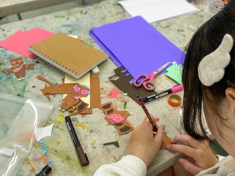 Image of A child with a white hair bow is making owl crafts with paper cut-outs, coloured markers, and glue at a cluttered art table scattered with notebooks, scissors, and coloured paper.