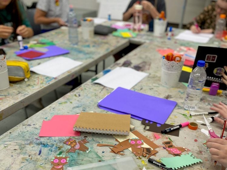 Image of A group of people sit around a paint-splattered table making crafts with colourful papers, notebooks, glue, and art supplies scattered across the surface. Some are assembling paper owls.