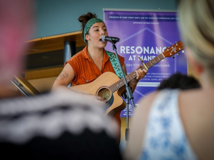 Image of A musician with a green headband and orange shirt sings and plays an acoustic guitar onstage, with a Resonance at Mana event banner in the background and blurred audience members in the foreground.