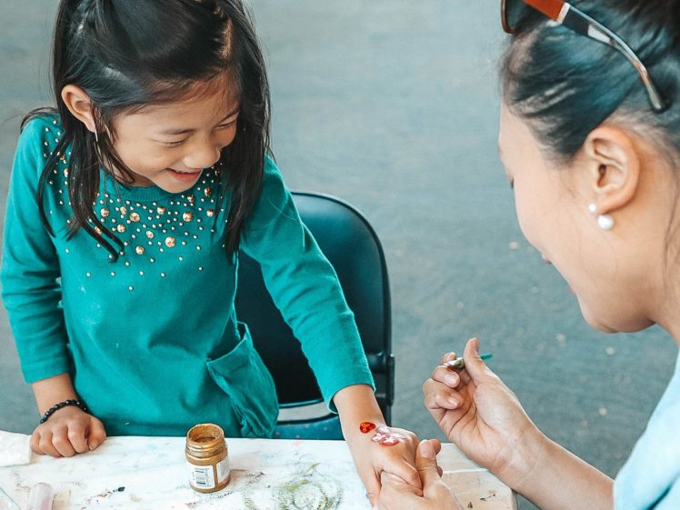 Image of A young girl smiles as an adult paints her hand with a brush at a table, with a small jar of paint nearby. The girl is wearing a teal shirt decorated with gold dots.