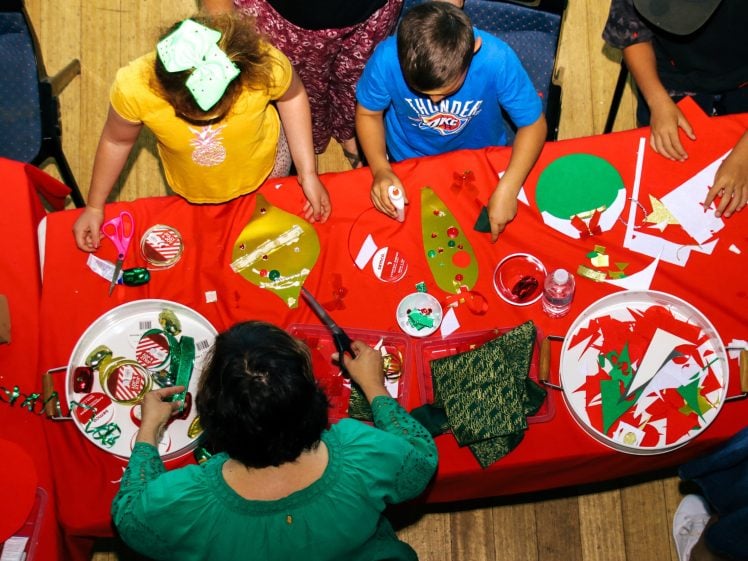 Image of Children and an adult gather around a red table, engaged in Christmas crafts with paper, scissors, and decorations. Cut-out shapes, ornaments, and craft supplies are scattered on trays and the tabletop.