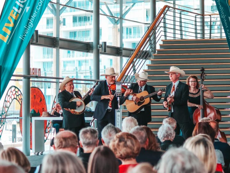 Image of A bluegrass band in cowboy hats performs with banjo, guitar, mandolin, and double bass on stage in front of a seated audience, inside a modern glass building with stairs and blue banners.