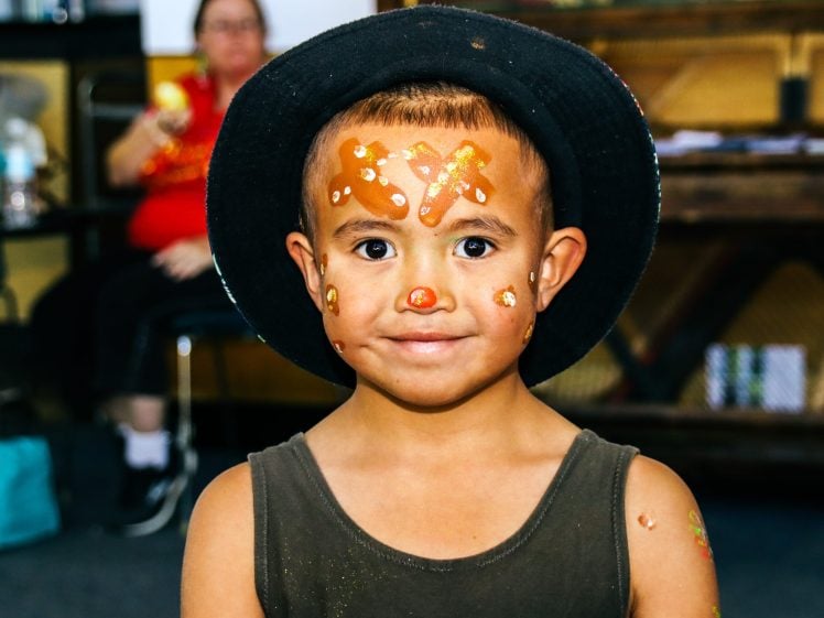 Image of A young child wearing a black hat and dark vest, with their face painted like a gingerbread man. The child is smiling, and there is a blurred adult figure in the background.