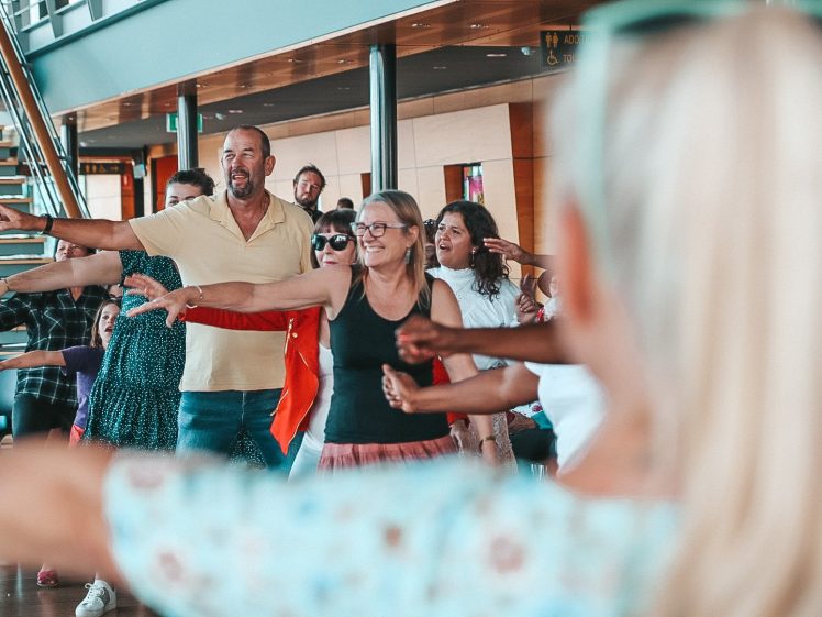 Image of A group of people smiling and standing with arms outstretched, participating in an indoor group activity or dance class. They appear to be enjoying themselves in a bright, modern space.