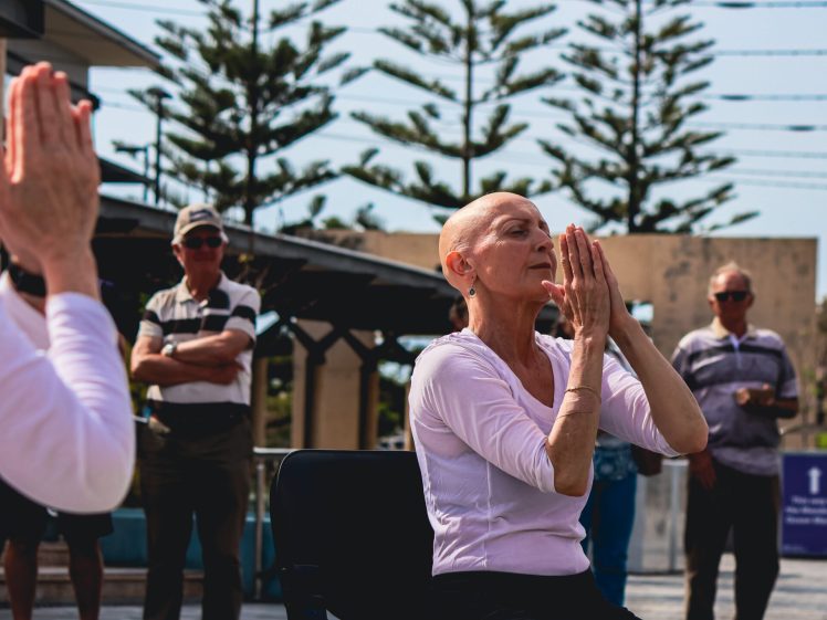 Image of An older woman sits outdoors with hands pressed together in a prayer pose, eyes closed, meditating. Several people stand in the background watching. Pine trees and fairy lights are visible behind them.