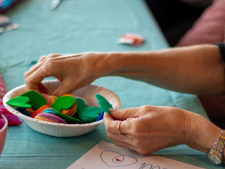 Image of A person’s hands sorting colourful felt hearts in a bowl on a teal tablecloth, with a flyer nearby that reads “1000 Hearts – a kindness project.” Other people and crafting supplies are in the background.