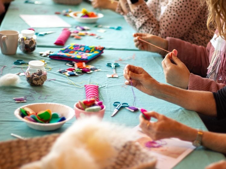 Image of People sitting at a table covered with craft supplies, including thread, scissors, felt shapes, buttons, and jars, working on creative projects with their hands.