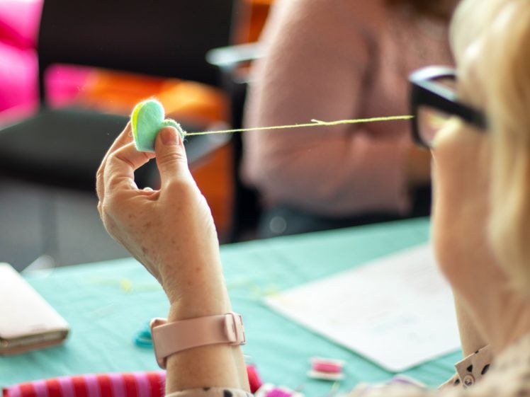 Image of A person wearing glasses and a pink watch holds a blue felt heart, threading a needle through it, with craft materials and blurred figures in the background.