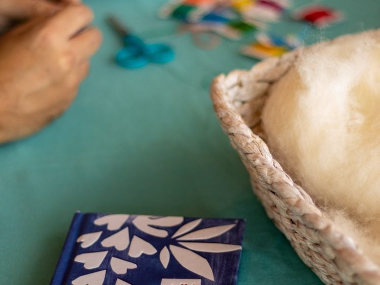 Image of A blue diary with white leaf designs and the words Love Your Life sits on a teal table near a woven basket of white wool, with crafting supplies and a person's hand in the background.