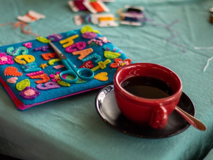 Image of A red mug of coffee sits on a black saucer with a spoon beside a colourful felt craft project, scissors, and embroidery threads on a teal fabric surface.