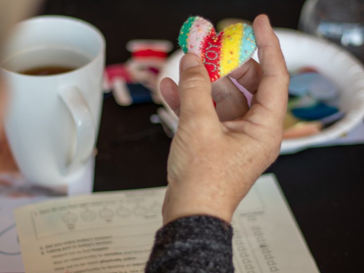 Image of A person holds a colourful, hand-sewn heart ornament above a table with a white mug, paperwork, and craft supplies in the background.