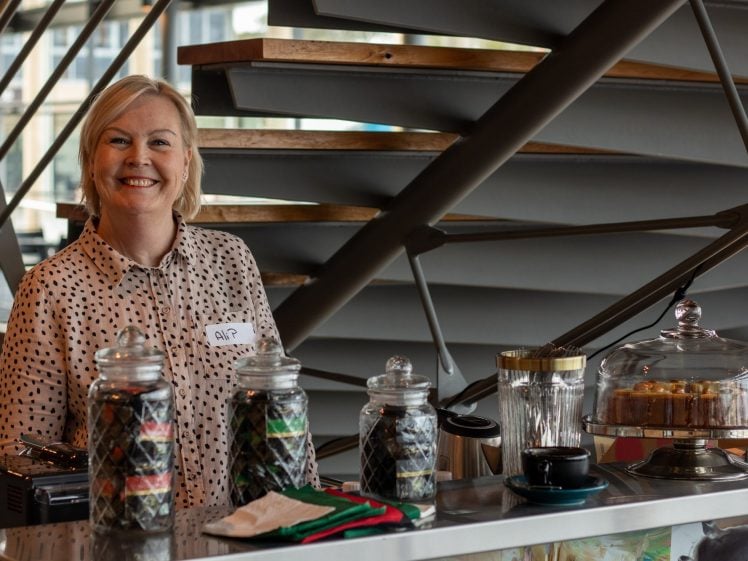 Image of A woman with short blonde hair, wearing a polka dot shirt and name badge, stands smiling behind a counter with jars of coffee pods, a glass cake stand with pastries, cups, and coffee equipment in a modern café setting.