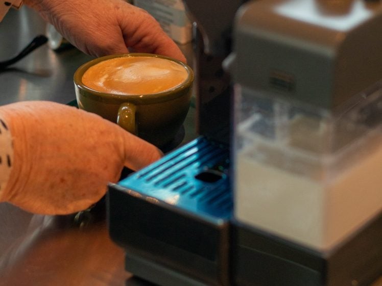 Image of A person with a polka-dot shirt holds a green mug of coffee with frothy milk beside an espresso machine on a kitchen worktop.