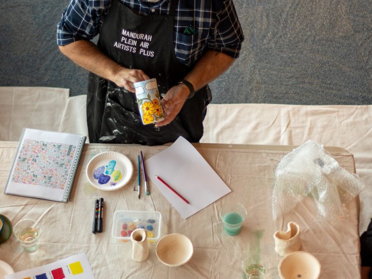 Image of A man in a black apron labelled Mandurah Plein Air Artists Plus paints a ceramic piece at a table covered with paint palettes, brushes, paper, and art supplies. The scene is viewed from above.