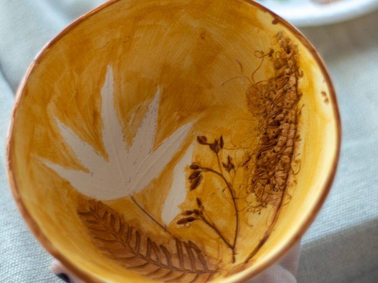 Image of A hand holds a yellow ceramic bowl decorated with painted botanical designs, including leaves and branches. In the background, a paper plate with various paint colours is visible on a light fabric surface.