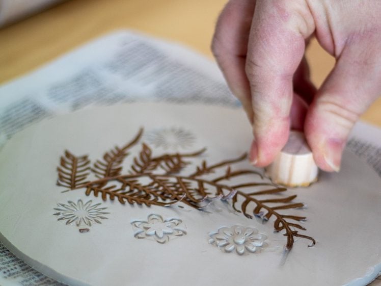 Image of A hand presses a small round stamp onto a flat piece of clay decorated with detailed fern leaves and flower designs. The clay sits on top of a newspaper.