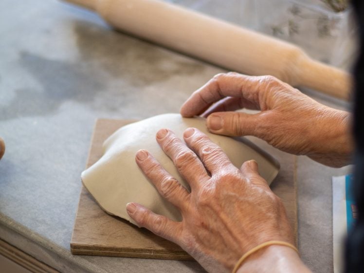 Image of A person shapes a piece of dough on a wooden board with both hands. A rolling pin and other kitchen items are visible on the worktop in the background.
