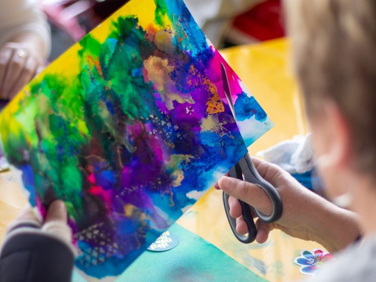 Image of A person uses scissors to cut a colourful, abstract painting on paper, featuring vibrant blue, green, yellow, and purple shades. Another hand is visible in the background on a yellow tabletop.