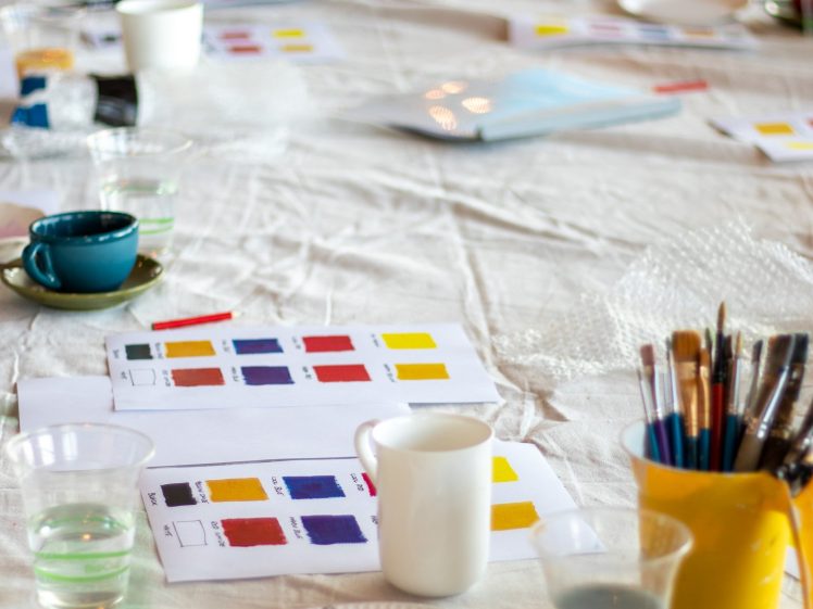 Image of A long table set up for painting, featuring colour swatches on paper, cups of water, paintbrushes in containers, and coffee cups. The table is covered with a light cloth, and people are visible in the background.