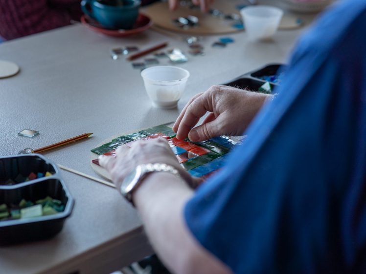 Image of A person arranges colourful mosaic tiles on a board at a table, surrounded by small containers and art supplies, with other people working nearby.
