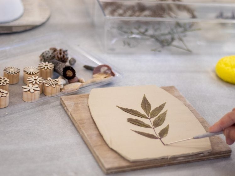 Image of A person uses a tool to press a real leaf into a slab of clay on a wooden board. Nearby are wooden stamps with floral patterns, natural objects, and a yellow sponge.
