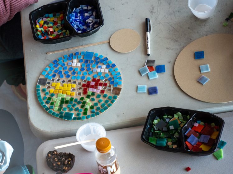 Image of A colourful mosaic art project in progress on a round base sits on a table, surrounded by small square tiles in various colours, containers, a marker, and a bottle of chocolate milk.