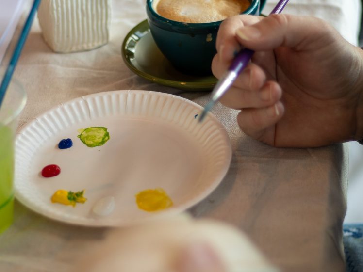 Image of A person holds a paintbrush over a paper plate with blobs of paint, next to a ceramic cup of coffee on a saucer and a white textured vase on a table covered with a cloth.