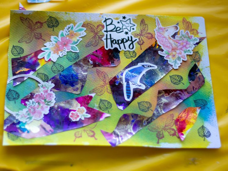 Image of A colourful handmade collage on paper with floral and bird stickers, painted abstract shapes, leaf and butterfly motifs, and a “Be Happy” cut-out on a yellow background.