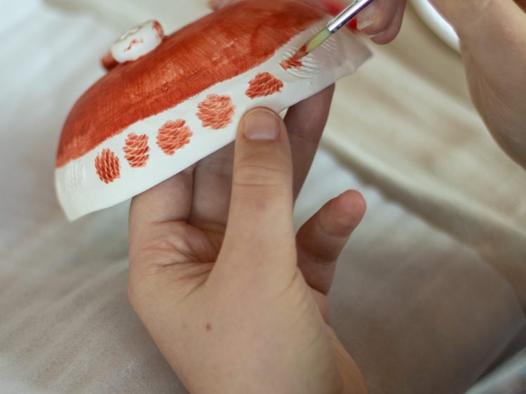 Image of A person uses a small paintbrush to paint red designs on a white ceramic dish, carefully adding decorative patterns around the edge.