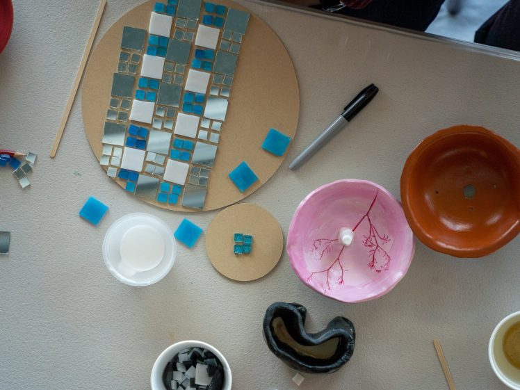 Image of A tabletop with mosaic-making supplies: a circular board with a blue and white tile mosaic, small tiles, a marker, bowls, a coffee cup, and craft tools arranged neatly.