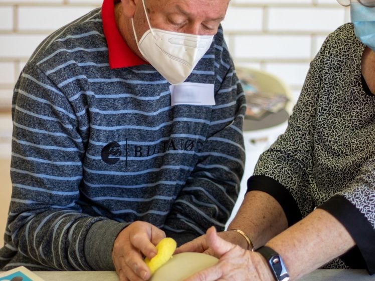 Image of Two older adults, both wearing face masks, work together shaping clay with their hands at a table. One person uses a yellow sponge, whilst the other guides their hands. A colourful sheet is visible on the table.