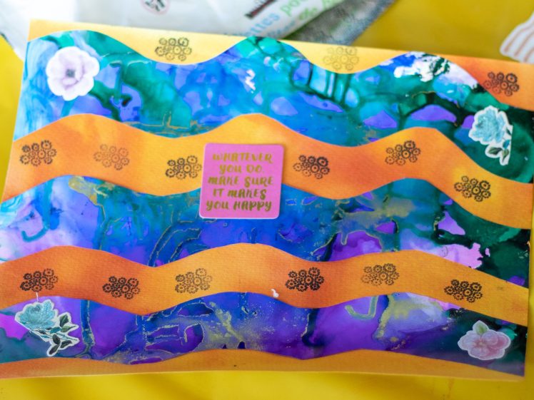 Image of A colourful art piece features wavy orange lines over a blue and purple background, with stickers of flowers and leaves. At the centre, a pink note reads, “WHATEVER YOU DO, MAKE SURE IT MAKES YOU HAPPY.”.