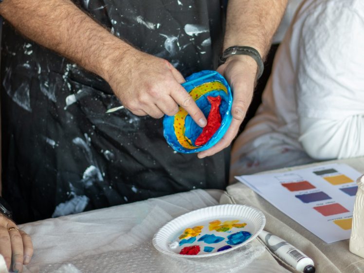 Image of A person wearing a paint-splattered apron holds a colourful, circular clay model, pointing at it. Paints on a paper plate and a colour chart are on the table in front of them.