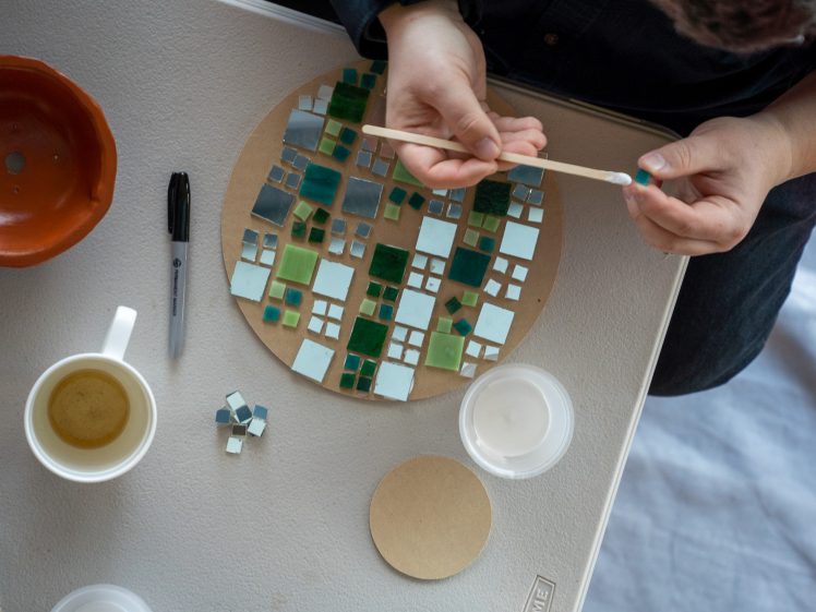Image of A person arranges and glues small green, blue, and white mosaic tiles onto a round board on a table. Nearby are a cup of tea, a marker, a bowl, adhesive, and a few loose tiles.