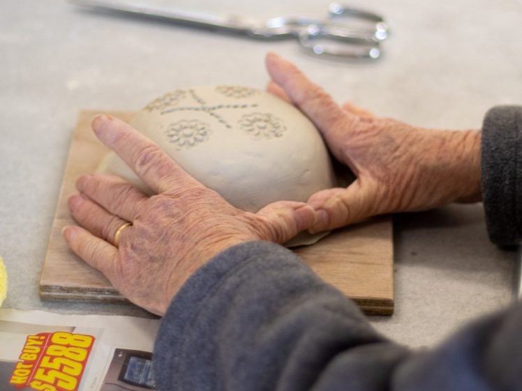 Image of A person with a gold ring shapes a piece of clay decorated with floral patterns on a wooden board. Scissors and a leaflet are on the table nearby.