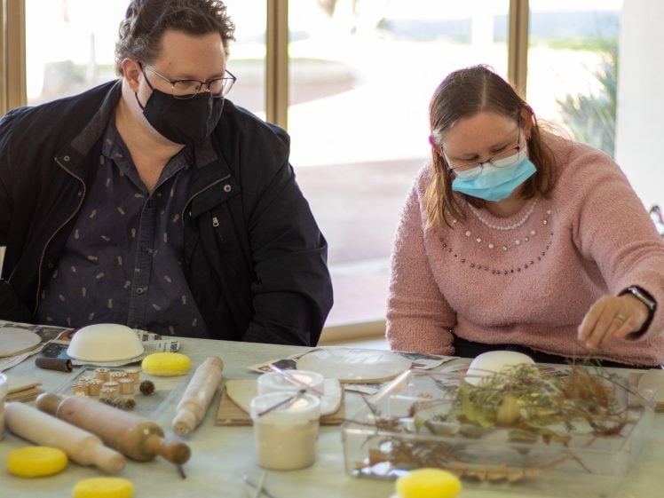 Image of Two people wearing masks sit at a table with art materials, including clay, rolling pins, sponges, and plants. The woman on the right arranges leaves, while the man on the left watches. They are indoors, near large windows.