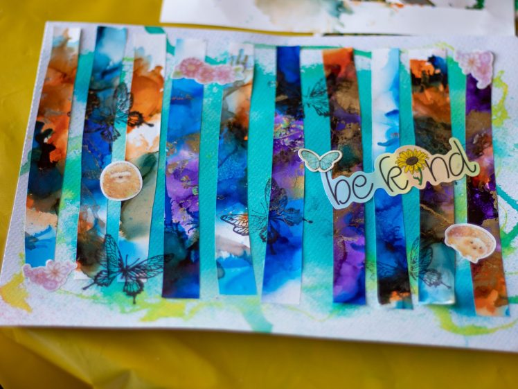 Image of A colourful mixed-media artwork with vertical blue and multicoloured strips, butterfly illustrations, flower stickers, and the words be kind with a sunflower, on a yellow surface.