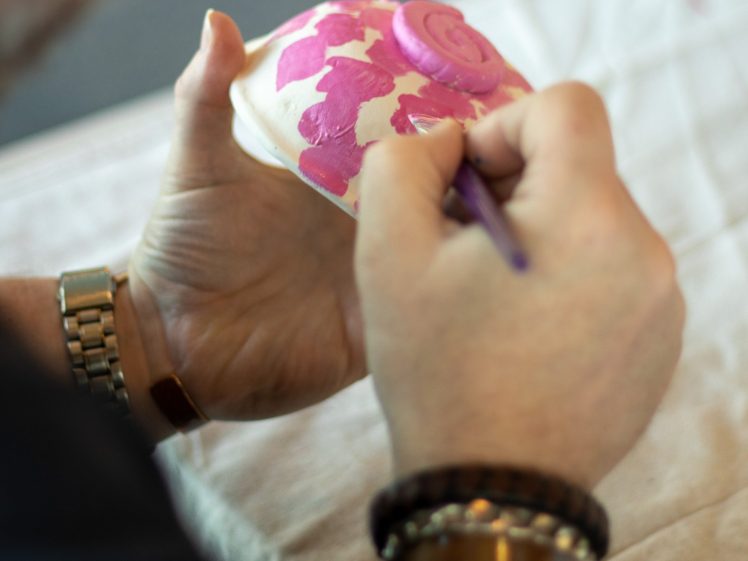 Image of A person wearing a watch and bracelets paints pink designs on a white ceramic object, possibly a cup or bowl, using a purple paintbrush. A beige cloth covers the table underneath.