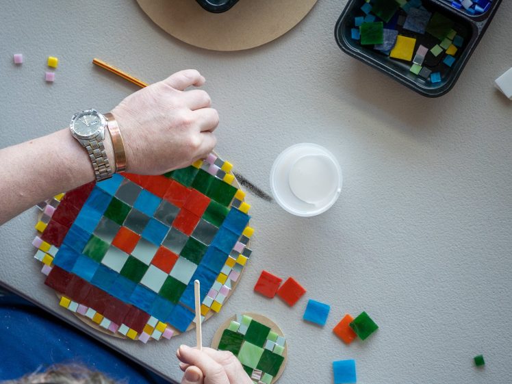 Image of A person arranges colourful square mosaic tiles onto a circular base, surrounded by loose tiles, a paintbrush, and a container of glue on a light surface.