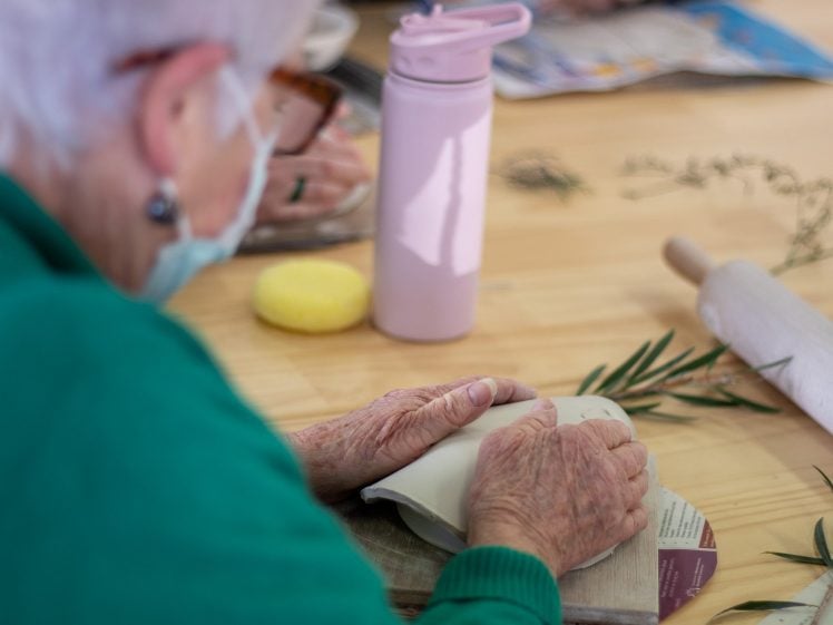 Image of An elderly person in a green jumper shapes clay with their hands on a wooden table. Nearby are a rolling pin, a yellow sponge, a pink water bottle, and leafy branches.