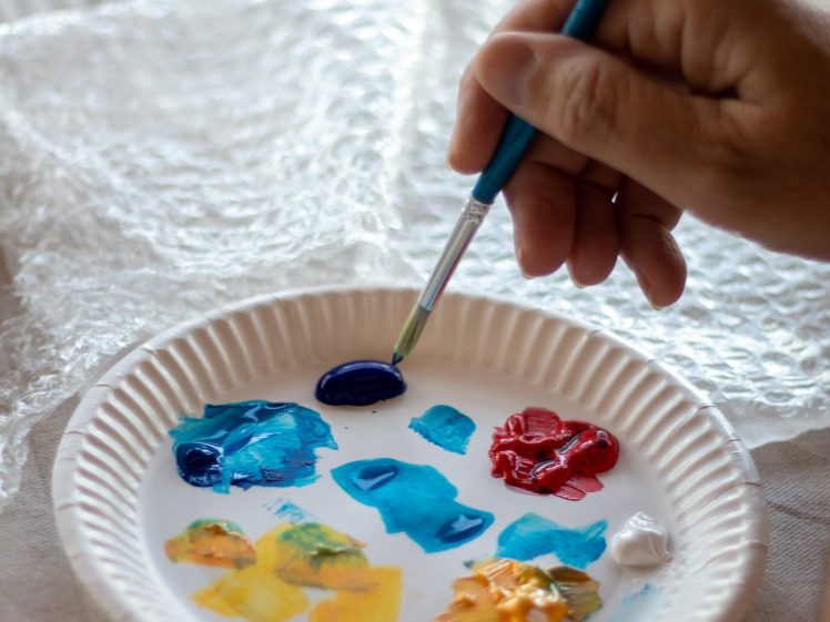 Image of A hand holds a paintbrush above a paper plate used as a palette, with dabs of blue, red, yellow, orange, and white paint on it. Bubble wrap is in the background.