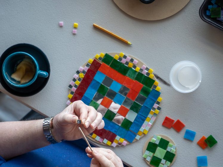 Image of A person is arranging colourful mosaic tiles on a round surface, with a pencil, cup of coffee, adhesive, and extra tiles nearby on the table. The person's hands are holding a tool, wearing a watch.